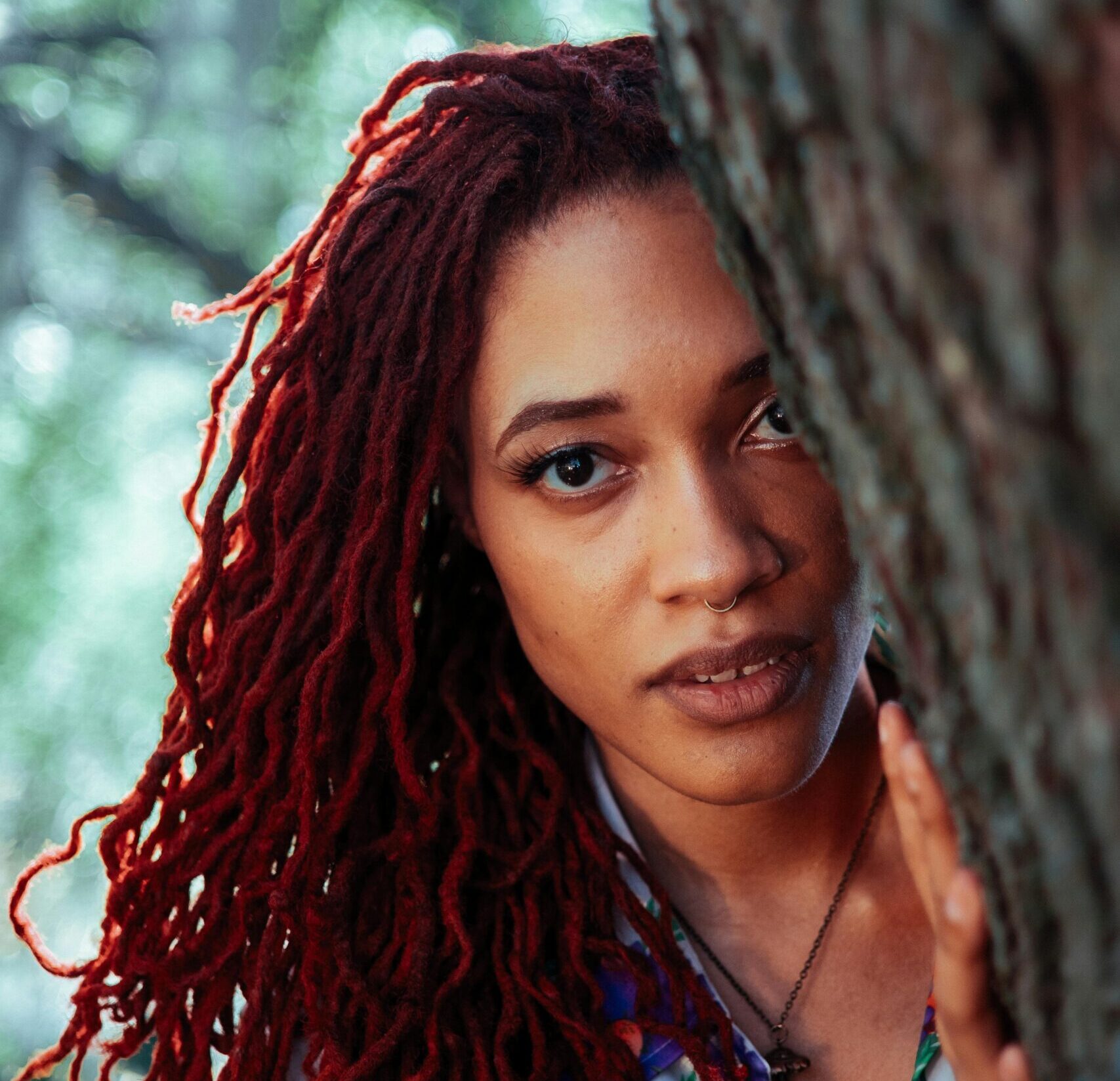 Beautiful portrait of a woman with red dreadlocks peeking from behind a tree outdoors.
